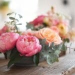 A rustic wooden table holds a low, round floral arrangement featuring vibrant pink peonies, soft orange roses, and eucalyptus leaves against a blurred