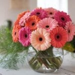 A vibrant bouquet of pink, orange, and peach Gerbera daisies is arranged in a clear glass fishbowl vase, surrounded by feathery green foliage, on a ru