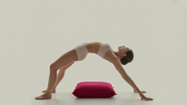 A woman in light activewear performs a yoga backbend, known as wheel pose, with her hands and feet on the floor and a pink cushion placed beneath her back. The background is plain and light-colored.