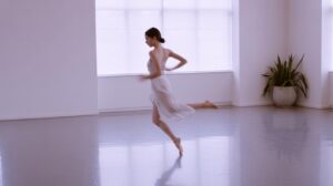A ballerina wearing a light, sheer dress dances gracefully in a bright, minimalist studio with large windows and a potted plant in the corner.