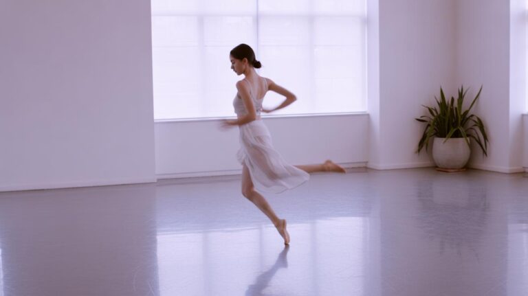 A ballerina wearing a light, sheer dress dances gracefully in a bright, minimalist studio with large windows and a potted plant in the corner.
