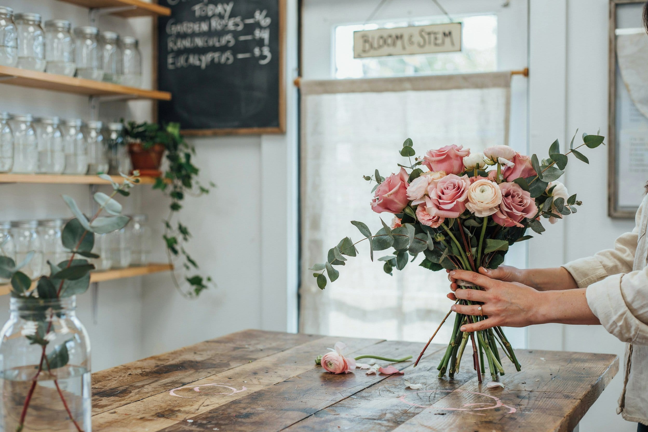 A florist arranges a fresh bouquet of pink garden roses and eucalyptus on a wooden table in warm natural light